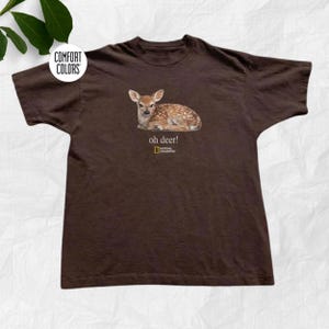 Könnte beinhalten: Ein dunkelbraunes T-Shirt mit kurzen Ärmeln und einer Grafik eines liegenden, gefleckten Rehkitzes. Unter dem Rehkitz steht der Text "oh deer!", gefolgt vom National Geographic Logo. Ein rundes Comfort Colors Markenetikett ist oben links zu sehen.
