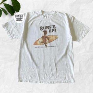 May include: A white short-sleeve t-shirt featuring a graphic of a brown monkey carrying a tan surfboard. Above the monkey, the text reads SURF'S UP! in dark brown letters. A circular logo in the top left corner contains the text COMFORT COLORS.