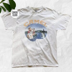 Könnte beinhalten: Ein weißes Kurzarm-T-Shirt mit einer Grafik eines Zeichentrick-Kamels, das in einem See angelt. Das Wort CAMEL ist in großen orangefarbenen Buchstaben über der Illustration gedruckt. Ein kleines kreisförmiges Logo mit dem Text COMFORT COLORS ist sichtbar.