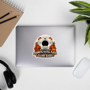 May include: A sticker featuring a medical CT scanner surrounded by autumn leaves and a pumpkin. The text below the scanner reads, "THANKFUL FOR FULL PLATES & CLEAR SLICES" in bold, dark lettering, set against a white border on a laptop surface.