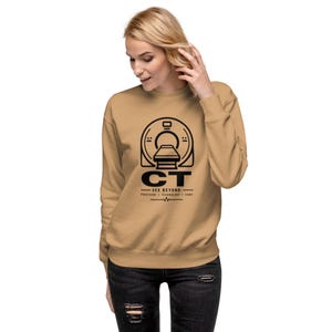 Peut inclure: Un sweat-shirt à col rond de couleur beige avec un motif noir représentant un scanner CT. Sous le scanner, le texte indique CT SEE BEYOND suivi des mots PRECISION TECHNOLOGY CARE et d'un petit graphique de ligne de rythme cardiaque.