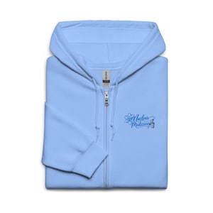 Puede incluir: Una sudadera con capucha y cremallera de color azul claro que presenta un diseño bordado en el pecho izquierdo. El bordado incluye un símbolo de átomo estilizado, una jeringa, un vial pequeño y el texto Nuclear Medicine en cursiva azul.