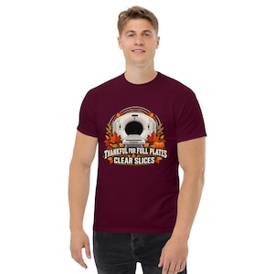 May include: A maroon short-sleeve t-shirt featuring a graphic of a CT scanner surrounded by autumn leaves and a pumpkin. The text reads, "THANKFUL FOR FULL PLATES & CLEAR SLICES" in a bold, stylized font centered below the medical imaging machine.