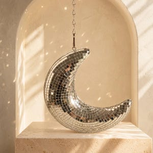 May include: A crescent moon-shaped disco ball covered in small, reflective silver mirror tiles. The object is suspended from a thin metal chain with a hook attachment, creating a curved, celestial form that catches and reflects light.