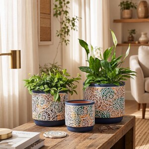 Op de afbeelding: Drie cilindervormige keramische plantenbakken met een donkerblauwe basis en rand, voorzien van een herhalend cirkelvormig patroon in tinten groenblauw, wit en koper. Twee plantenbakken bevatten groene planten, terwijl er één leeg is.