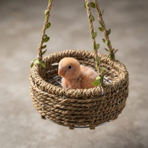 May include: A small, light orange baby bird with soft downy feathers sits inside a round, woven rope basket. The basket features a metal wire base and is suspended by two thick, braided rope handles adorned with small, green artificial leaves.