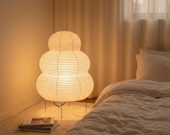 May include: A floor lamp with a three-tiered, rounded paper shade featuring horizontal wire ribbing. The lamp emits a warm, soft glow and is supported by thin, black metal legs. The structure is composed of three stacked, bulbous sections.