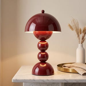 May include: A glossy dark red table lamp featuring a large dome-shaped shade positioned above a vertical stack of three spherical elements of varying sizes, all supported by a rounded base, creating a sculptural silhouette with a smooth finish.