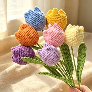 May include: A bouquet of seven crocheted tulips in various colors including blue, orange, purple, yellow, pink, and light yellow. Each flower features a textured yarn construction with green crocheted stems and long, pointed green leaves.