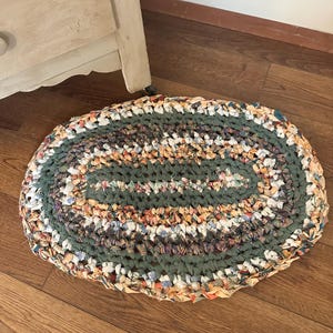 May include: An oval-shaped rag rug constructed from braided fabric strips in a mix of muted green, orange, cream, and dark brown tones. The rug features a concentric pattern of woven textile loops, creating a textured surface with varied colors.