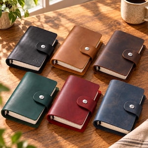 May include: Six small leather journals with snap closures arranged on a wooden surface. The journals come in black, tan, dark brown, dark green, red, and navy blue. The black journal features the name ZOE embossed on the front cover.