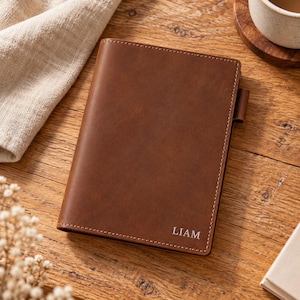 Pode incluir: Uma capa de caderno em couro castanho com acabamento liso e mate e costuras visíveis ao longo das bordas. O nome LIAM está gravado numa fonte sem serifa clara no canto inferior direito. Um pequeno suporte de couro para caneta está fixado na lateral.