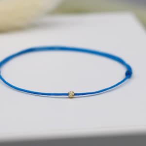 May include: A minimalist blue bracelet with a small, gold-coloured bead at the centre. The bracelet is made of a thin, bright blue cord. The bead appears to be approximately 0.25 cm in diameter. The bracelet is displayed on a white surface.