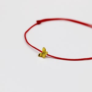 May include: A delicate red string bracelet featuring a small, gold-colored butterfly charm. The bracelet is simple and elegant, with a minimalist design. The butterfly charm adds a touch of whimsy to the jewelry.
