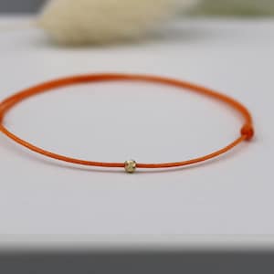 Könnte beinhalten: Orangenes Schnurarmband mit einer kleinen goldenen Perle in der Mitte. Das Armband hat ein minimalistisches Design mit einer leuchtend orangefarbenen Kordel. Die goldene Perle verleiht dem Design einen Hauch von Eleganz.