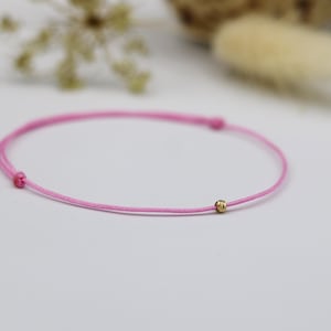 May include: A delicate pink bracelet with a small gold bead. The bracelet is made of a thin, pink cord and features a simple design. The gold bead is centered on the bracelet. The bracelet is a minimalist piece of jewellery.