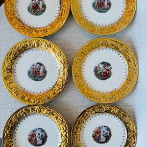 May include: A set of six round porcelain plates featuring a wide, embossed gold rim with intricate floral and vine detailing. Each plate has a central circular medallion depicting a colorful scene of figures in classical attire in a landscape.