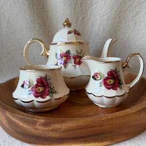 May include: A cream-colored ceramic tea set consisting of a teapot with a lid, a creamer, and a sugar bowl. Each piece features a floral pattern of dark pink camellia flowers with green leaves and is accented with gold trim on the edges.