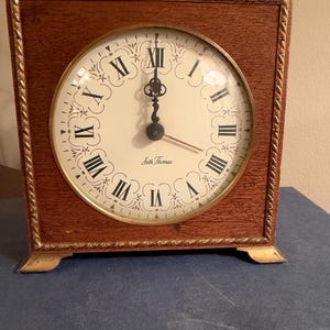 May include: A square wooden mantel clock with a gold-toned rope-style border and feet. The off-white clock face features black Roman numerals, decorative scrollwork, black hands, and the text Seth Thomas printed in the center.