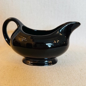 May include: A glossy black ceramic gravy boat featuring a curved handle, a wide pouring spout, and a rounded body that sits on a circular pedestal base. The surface has a smooth, reflective finish that catches light across its curved form.