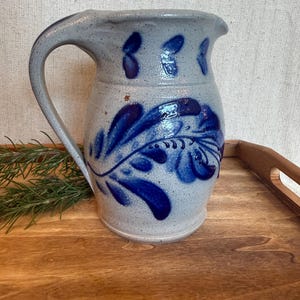 May include: A light grey speckled ceramic pitcher featuring a dark blue hand-painted floral leaf pattern. The pitcher has a curved handle on the left side, a wide spout, and a smooth glazed finish. The design includes small blue petal accents.