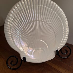 May include: A white ceramic dish shaped like a large scallop shell with a glossy, iridescent finish and radiating ridges. The dish is held upright by a black metal stand with two curved, scroll-like feet that support the base of the shell.