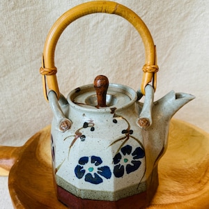 May include: A ceramic teapot with a speckled light gray body, a dark brown base, and a curved bamboo handle. The teapot features hand-painted blue floral motifs with green centers and a small brown knob on the lid. It has a faceted hexagonal shape.