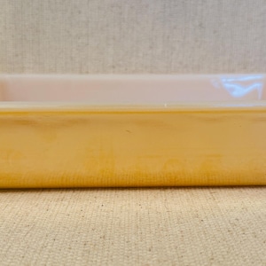 May include: A rectangular baking dish made of peach-coloured glass with a glossy, iridescent finish. The dish features smooth, rounded corners and a wide, flat rim, showcasing a translucent quality that allows light to pass through the material.