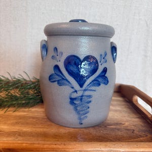May include: A light grey stoneware crock with a matching lid and two side handles. The front features a hand-painted cobalt blue heart design with decorative leaf-like accents and a swirling base pattern, finished with a glossy glaze.