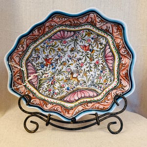 May include: A scalloped ceramic bowl featuring a white interior with intricate hand-painted floral patterns, small birds, and a deer. The rim is decorated with orange, pink, and teal accents. The bowl rests on a black metal display stand.