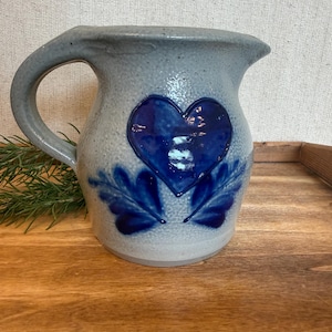 May include: A light gray speckled ceramic pitcher featuring a prominent, glossy dark blue heart design on the front, accented by two symmetrical blue leaf motifs below the heart. The pitcher has a curved handle and a wide, flared pouring spout.