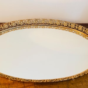 Puede incluir: Una bandeja ovalada con una base de espejo reflectante y un borde decorativo de metal dorado. El borde elevado presenta un intrincado patrón de hojas repetitivo a lo largo de todo el contorno, creando un marco texturizado para el centro liso.