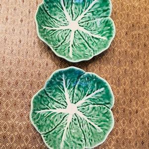 May include: Two ceramic dishes shaped like green cabbage leaves. Each dish features a glossy finish with realistic embossed textures, including prominent white veins radiating from the center to the scalloped, wavy edges of the leaf-shaped bowls.