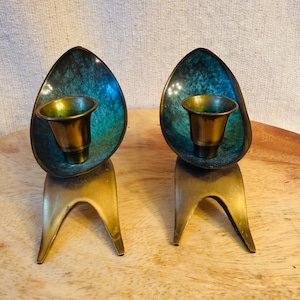 May include: A pair of brass candle holders featuring a tripod base and a central cup. Each holder has a curved, petal-like back panel with a mottled teal and green patina finish, contrasting with the polished brass of the cup and base structure.