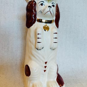 May include: A ceramic figurine of a dog standing on its hind legs. The dog is primarily white with brown patches on its ears and sides. It wears a gold-colored collar with a gold tag. There are three small red hearts on its chest and green leaf-like details at the top.