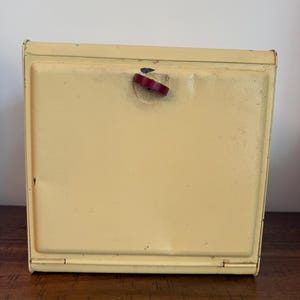 May include: A square, pale yellow metal box with a hinged front panel. The front features a small, dark red, semi-circular latch handle centered near the top edge. The metal surface shows minor signs of wear, including small scratches and paint chips.