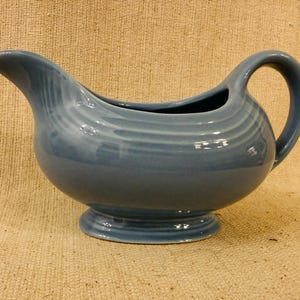 Peut inclure: Une saucière en céramique bleu poussiéreux avec une finition lisse et brillante, un large bec verseur, une anse incurvée et une base circulaire sur pied. La surface extérieure est ornée de subtiles rainures circulaires horizontales.
