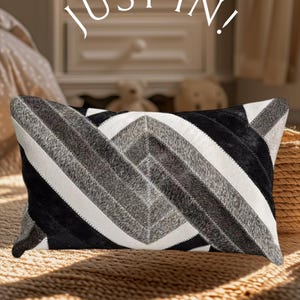 May include: A rectangular throw pillow featuring a geometric pattern of black, white, and gray hair-on-hide panels arranged in chevron stripes. The text "JUST IN!" is printed in white in the upper center of the image above the pillow.
