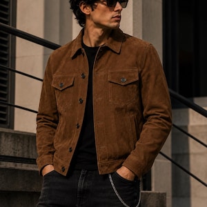 May include: A brown suede jacket featuring a classic collar, button-front closure, and two chest pockets with buttoned flaps. The jacket is worn over a black shirt and paired with dark denim jeans that include a silver chain accessory at the waist.