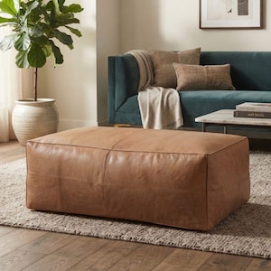 May include: A rectangular tan leather ottoman with visible stitched seams along the top and sides. The ottoman has a smooth, matte finish and a low-profile design, positioned on a textured rug in front of a dark teal sofa with decorative pillows.