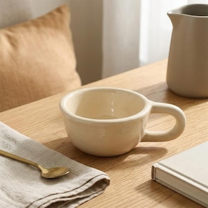Pode incluir: Uma caneca de cerâmica na cor creme com uma tigela larga e arredondada e uma alça grossa e curva. A caneca tem um acabamento liso e brilhante com um design simples e funcional. Está posicionada sobre uma superfície de madeira.