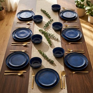 Puede incluir: Una mesa de comedor preparada para seis personas con platos y cuencos de cerámica azul oscuro moteado, acompañados de cubiertos dorados que incluyen tenedores, cuchillos y cucharas. Un camino de mesa de lino beige está decorado con ramas de eucalipto.