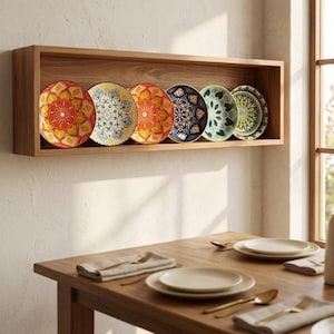 May include: A rectangular wooden wall shelf holding six decorative plates with intricate, colourful mandala-style patterns in shades of orange, blue, yellow, and green, positioned above a wooden dining table with place settings.