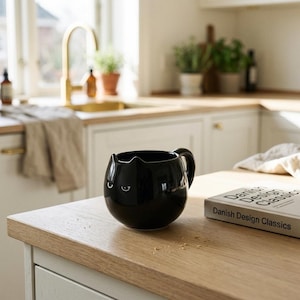 May include: A glossy black ceramic mug shaped like a round cat head with small pointed ears and a simple white graphic of two eyes. The mug features a curved handle on the right side and is positioned next to a book titled Danish Design Classics.