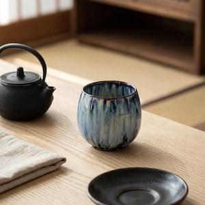 Könnte beinhalten: Eine abgerundete Keramik-Teetasse mit einer reaktiven Glasur in tiefblauen, weißen und dunkelbraunen Streifen. Die Tasse steht neben einer schwarzen gusseisernen Teekanne und einer passenden schwarzen Untertasse auf einer hellen Holzoberfläche.