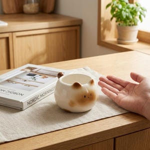 Może przedstawiać: Okrągły, kremowy kubek ceramiczny w kształcie kota, z brązowymi końcówkami uszu, małym brązowym ogonkiem pełniącym funkcję uchwytu oraz prostym wyrazem twarzy z dwojgiem czarnych oczu i maleńkimi ustami, umieszczony obok książki.