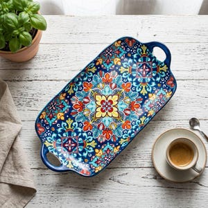 May include: A rectangular ceramic serving tray with two integrated handles, featuring a vibrant, symmetrical mandala-style pattern in shades of deep blue, bright yellow, orange, and turquoise, with a glossy finish and curved edges.