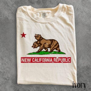 May include: An ivory-coloured t-shirt featuring a graphic of a two-headed brown bear standing on a green patch of grass. To the left is a red star. Below the bear is a red rectangular bar with the white text NEW CALIFORNIA REPUBLIC.