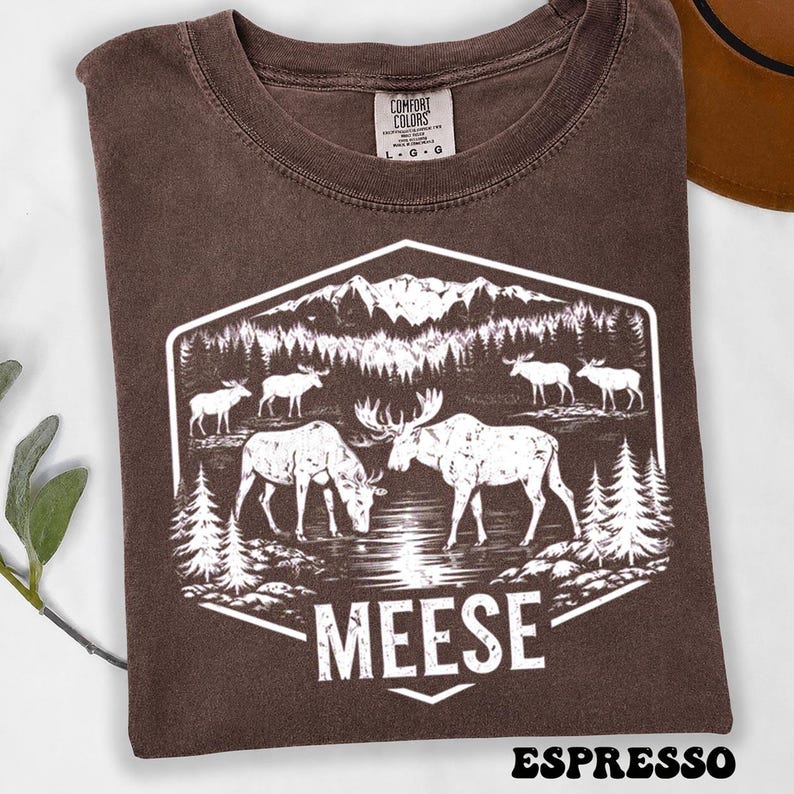 May include: A brown t-shirt featuring a white graphic print of a mountain landscape with pine trees and several moose standing near a body of water. The text MEESE is printed in white at the bottom of the hexagonal design. The shirt has a Comfort Colors label.