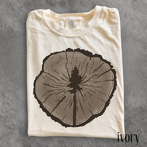 May include: An ivory-colored short-sleeve t-shirt featuring a dark brown graphic of a tree stump cross-section with a pine tree silhouette in the center. The inner neck label displays the text Comfort Colors. The fabric has a visible texture.